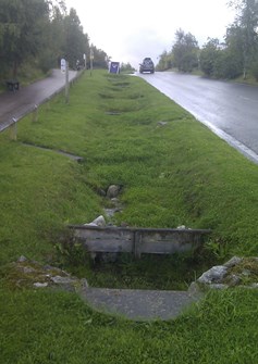 Grassy swale next to steep road with check dams to slow flow of water